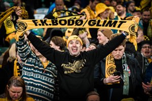 En Glimt-supporter med gul lue fra J-felet, svart genser fra J-feltet holder et gult skjerf med teksten "FK Bodø/Glimt" over hodet på bortefeltet på Celtic Park i Glasgow i februar 2022