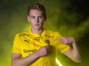 Isak Dybvik Määttä peker på Glimt-logoen på drakten han har på seg foran gule lys og en røyksky