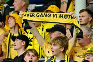 Gule Glimt-supportere på tribunen på Aspmyra under oppgjøret mot RFS i 2024. I midten av bildet holder en mann med svart caps opp et skjerf med teksten "Hver gang vi møtes"