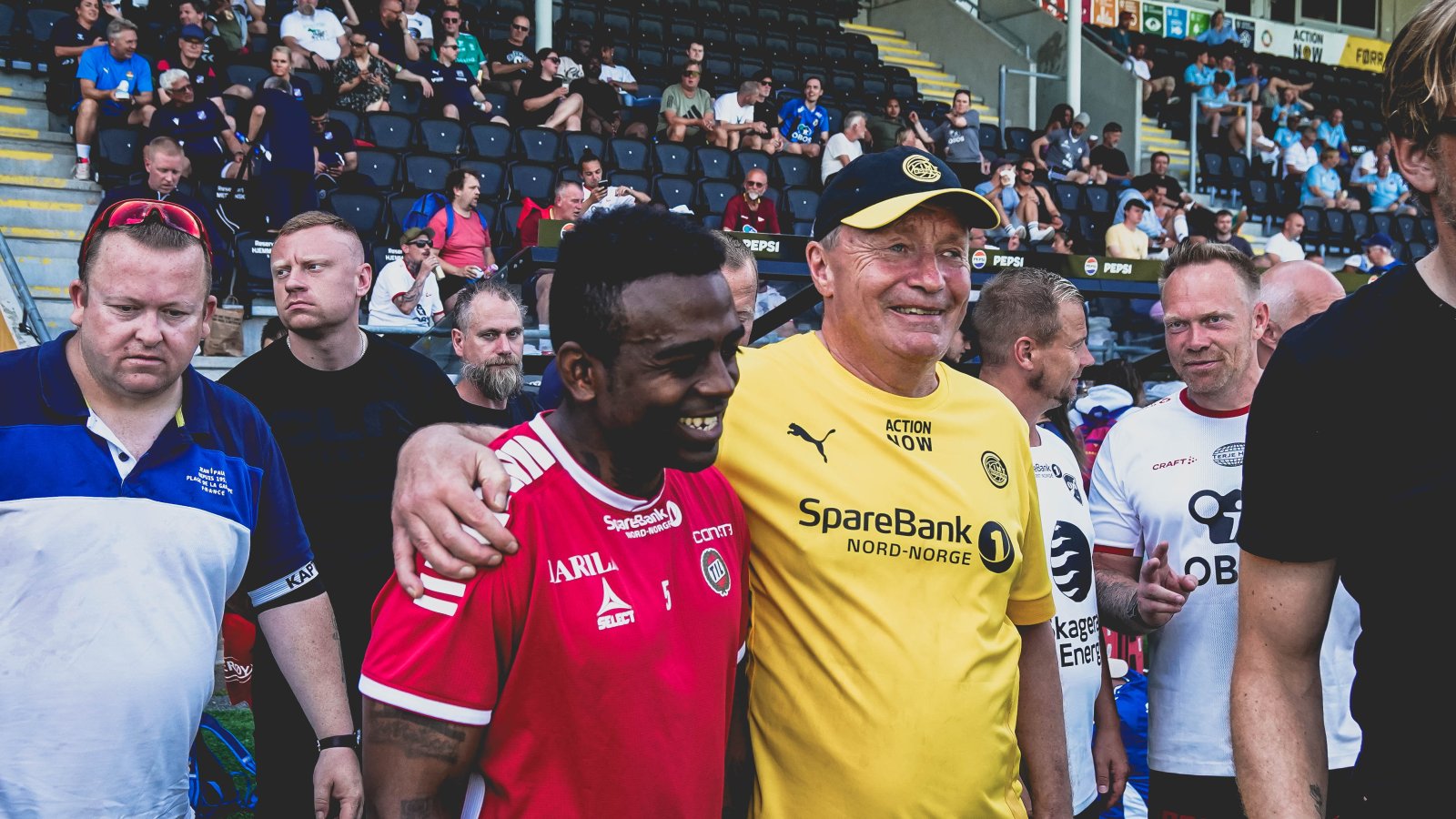 En smilende Glimtvis-spiller med Bodø/Glimt-drakt og Bodø/Glimt-caps holder rundt en TIL-spiller på Aspmyra stadion i Bodø i juni 2024 mens spillere og trenere fra andre lag virmler rundt under NM i gatefotball 2024