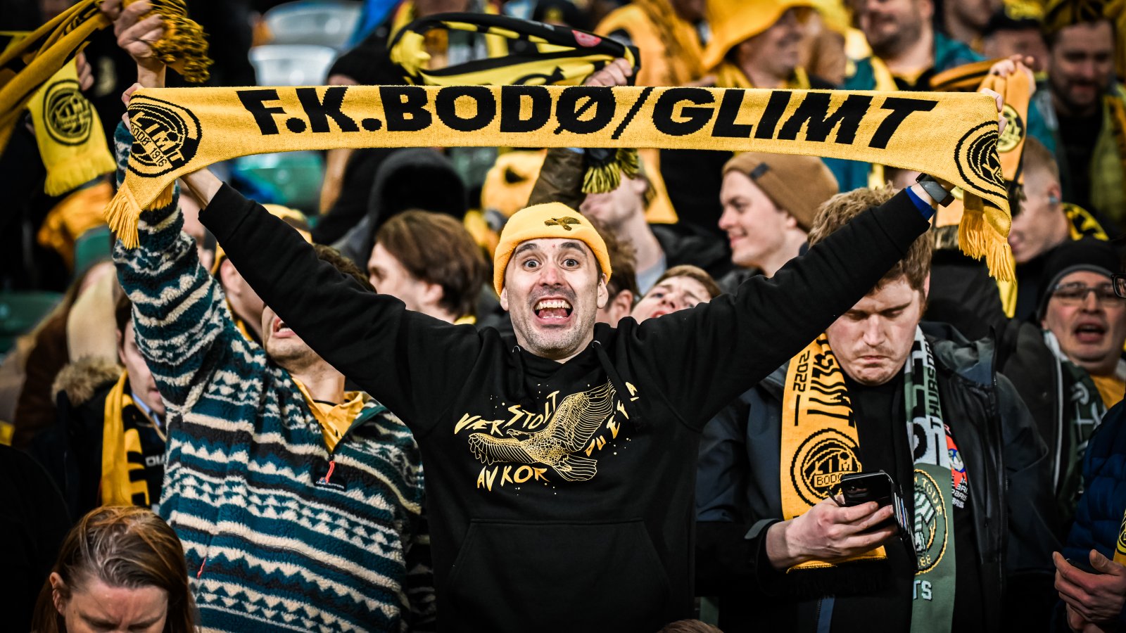 En Glimt-supporter med gul lue fra J-felet, svart genser fra J-feltet holder et gult skjerf med teksten "FK Bodø/Glimt" over hodet på bortefeltet på Celtic Park i Glasgow i februar 2022