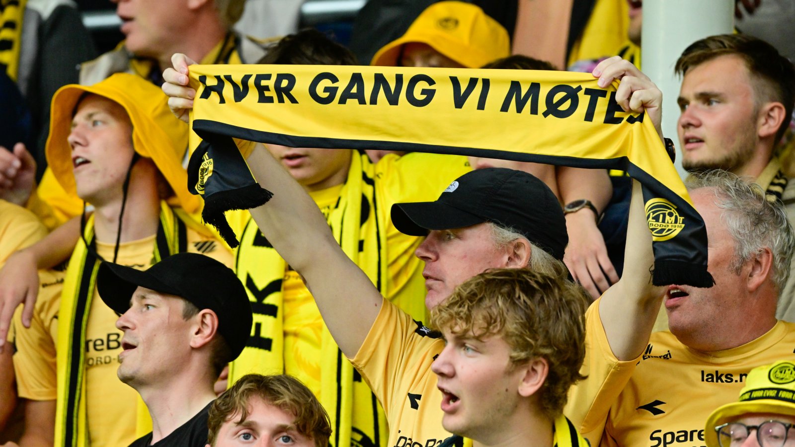 Gule Glimt-supportere på tribunen på Aspmyra under oppgjøret mot RFS i 2024. I midten av bildet holder en mann med svart caps opp et skjerf med teksten "Hver gang vi møtes"